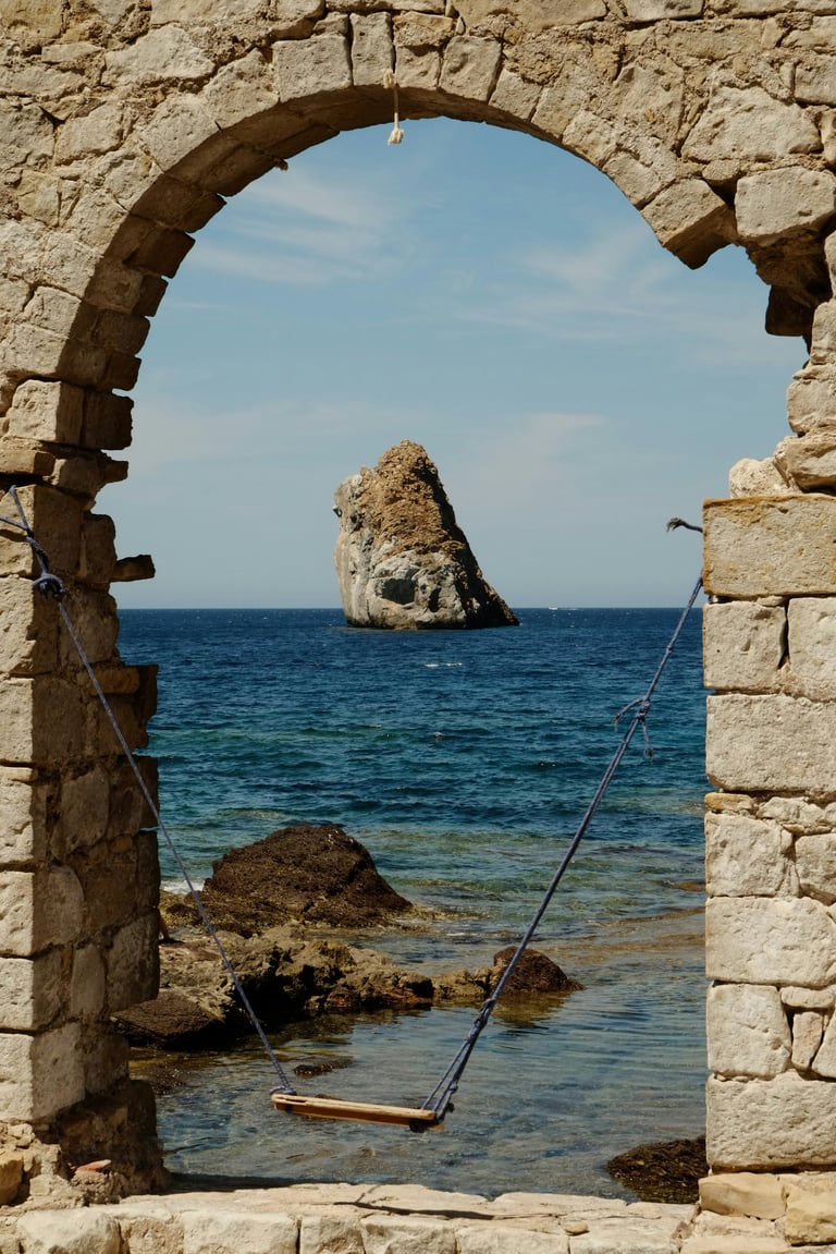 Distant rock formation visible through a stone archway opening to a Mediterranean sea with a rope swing in the foreground