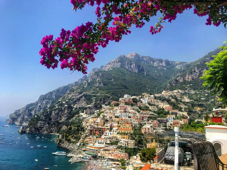 Vibrant purple bougainvillea flowers frame a picturesque coastal village built on steep cliffs overlooking the turquoise Mediterranean Sea