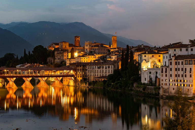 Historic riverside town with illuminated medieval buildings and stone bridge reflected in calm water at dusk, mountains in background